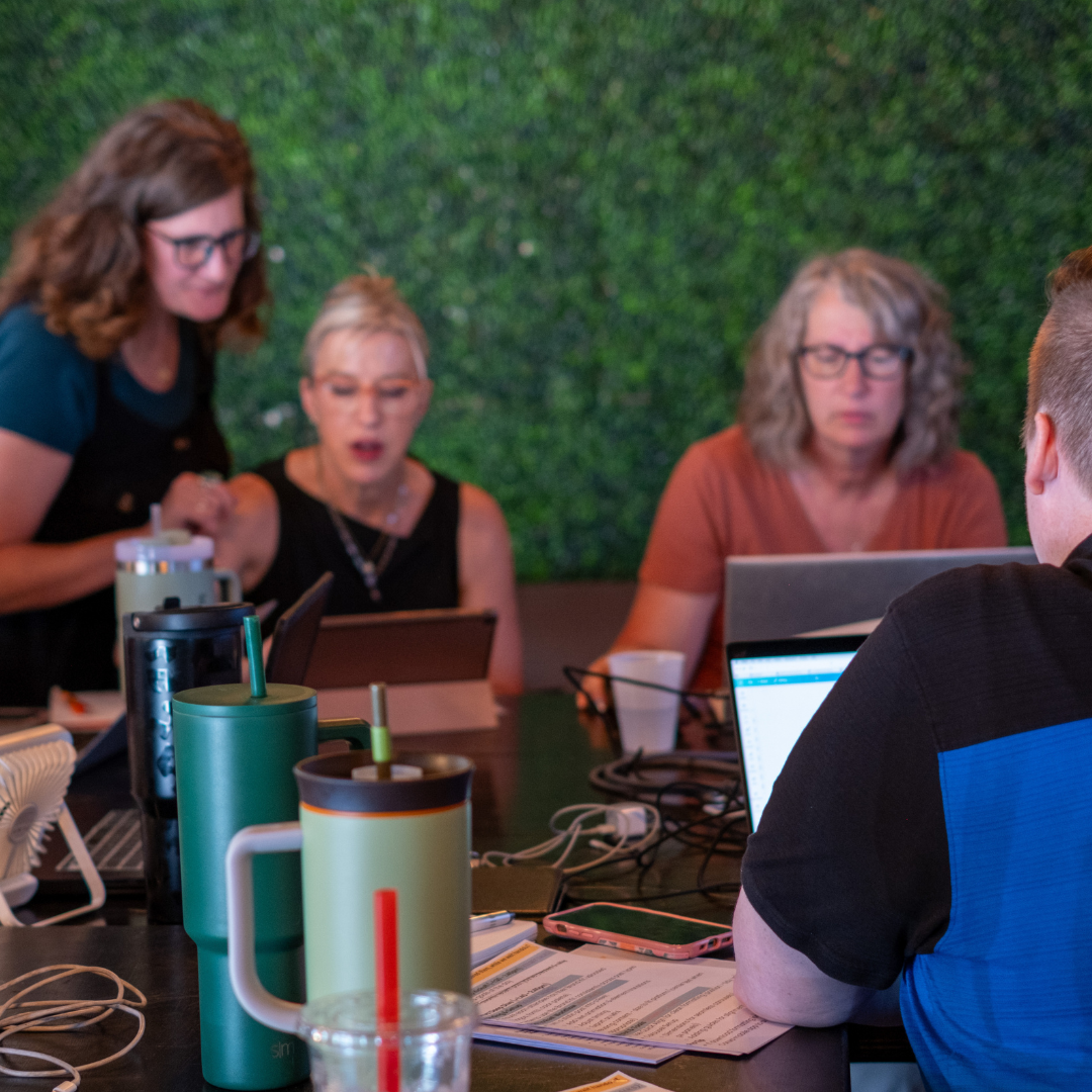 Group of people working together at a table with laptops and coffee mugs in front of a green wall.